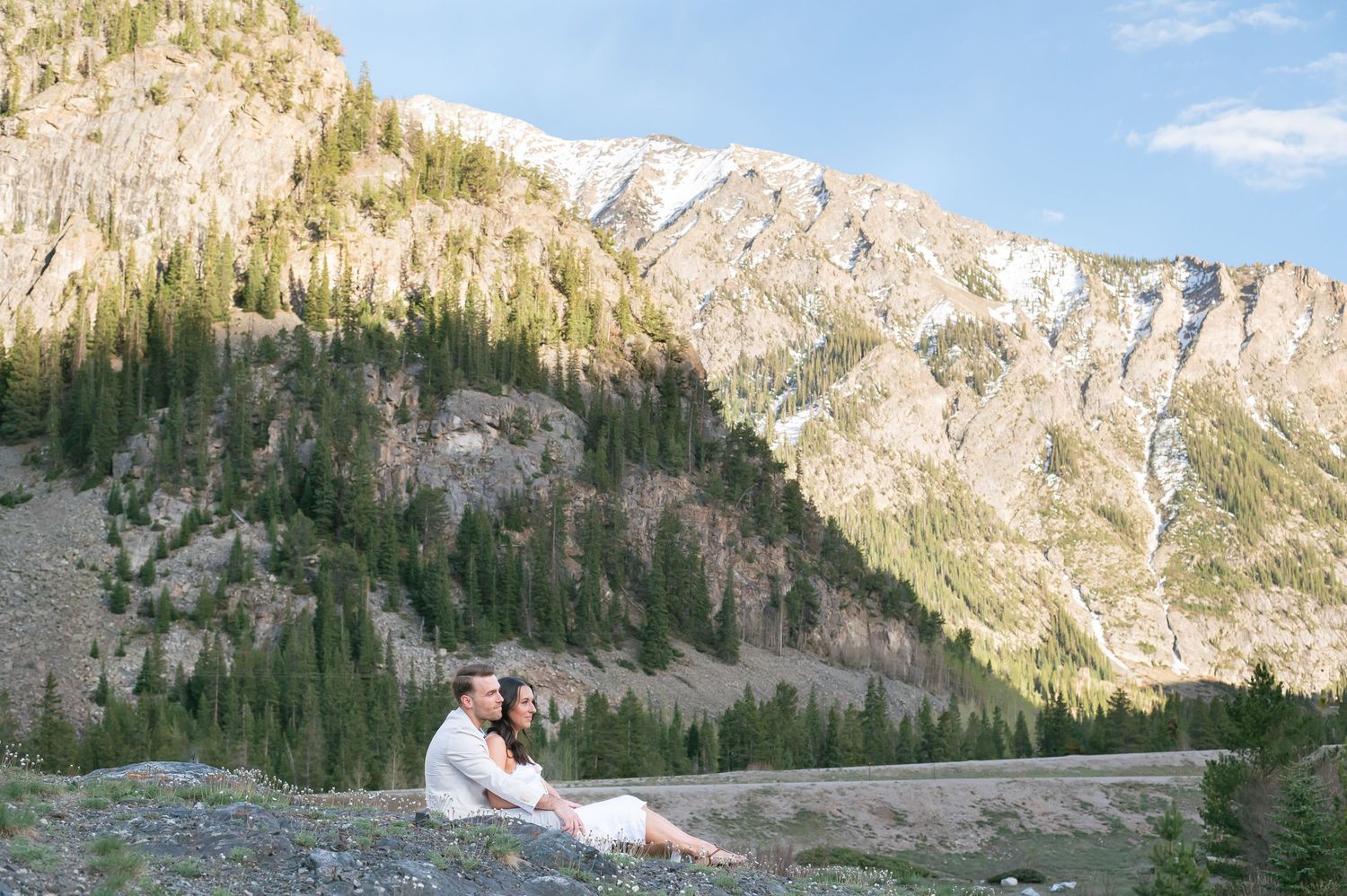 Couple sitting together on rocky mountainside surrounded snow-capped peaks in golden hour light for an elopement at Officer's Gulch