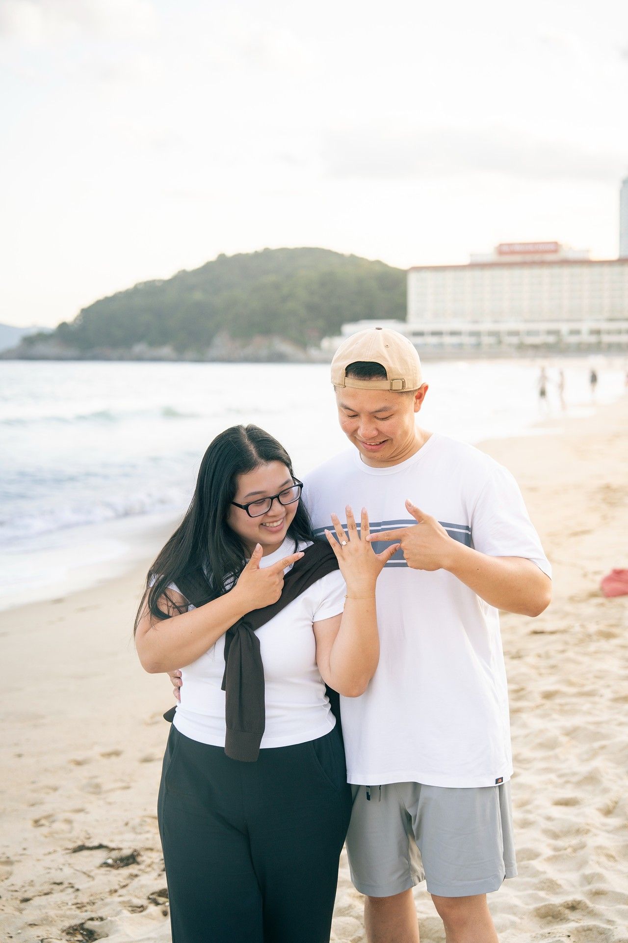 Couple on beach admiring engagement ring, smiling joyfully, with ocean waves and resort building in background.
