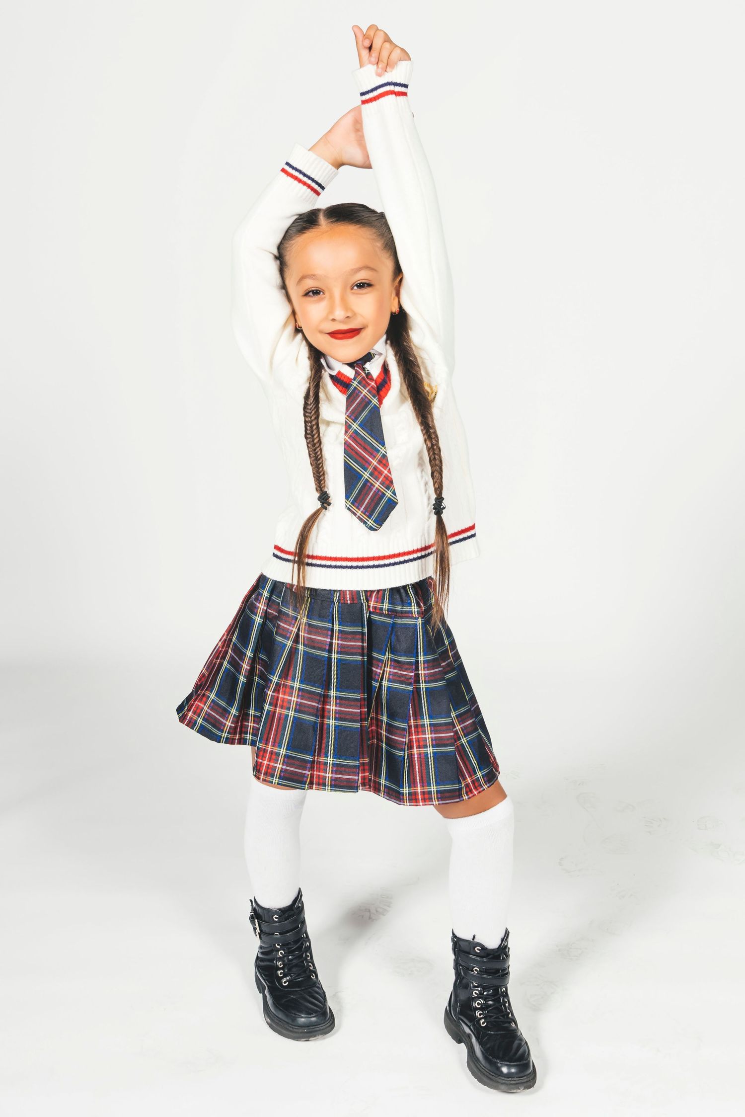 Young girl in plaid school uniform skirt, white sweater, tie, and black boots poses with arms raised.