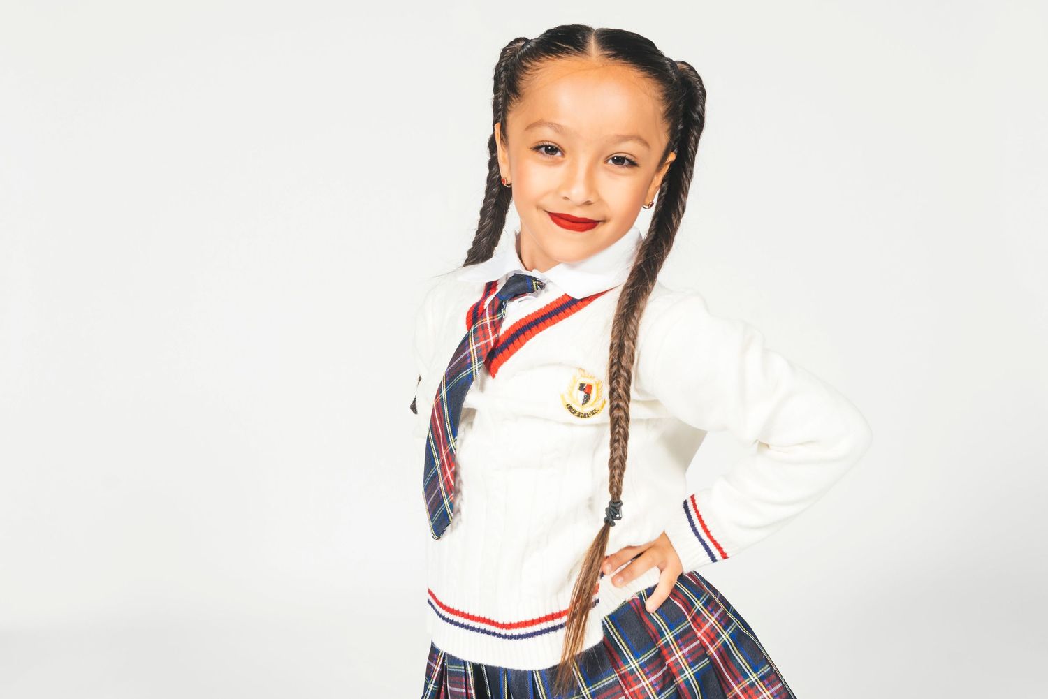 Young girl in school uniform with braids, tie, and plaid skirt poses confidently on white background.