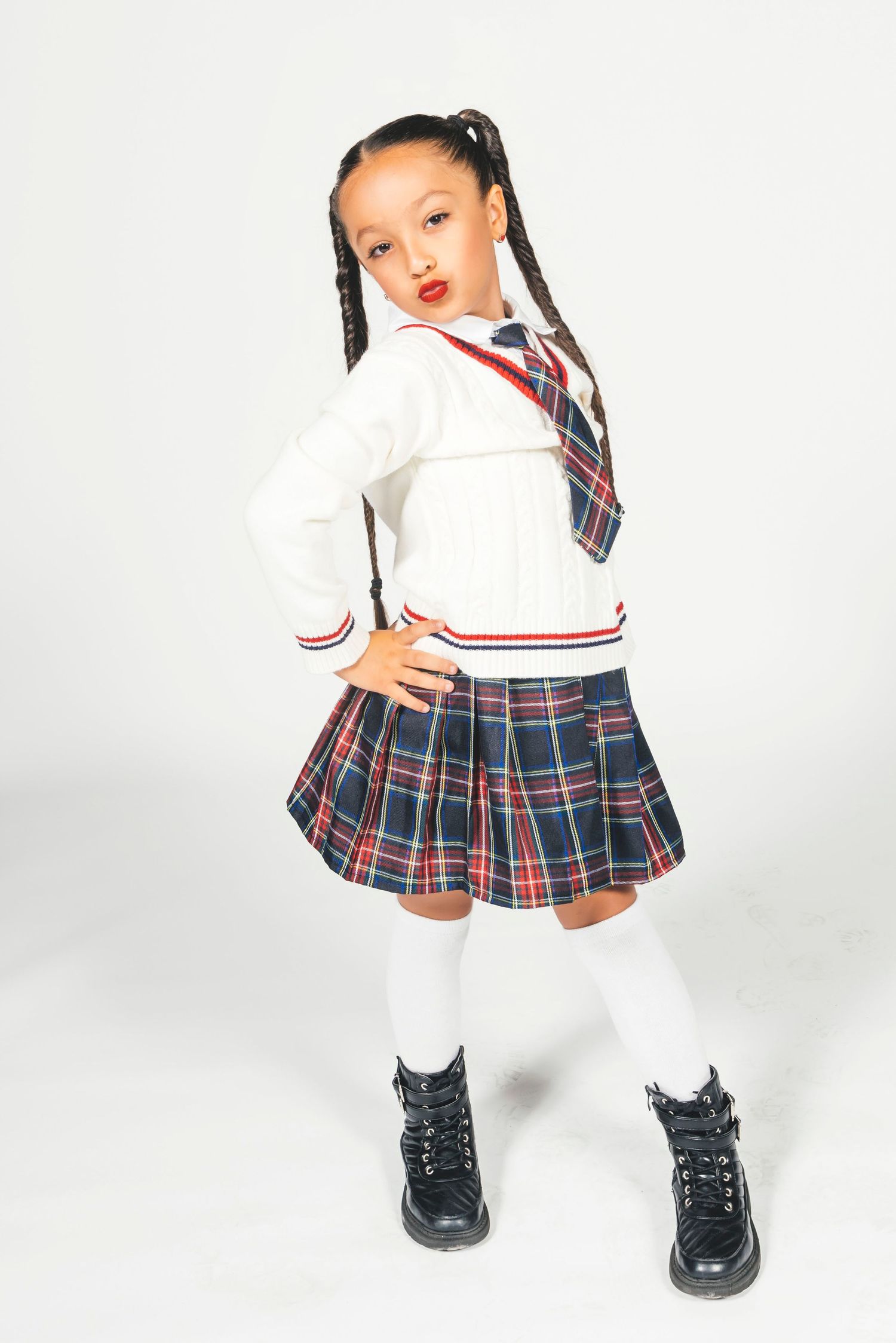 Young girl in school uniform with plaid skirt, white sweater, tie, knee socks, and black boots, posing confidently.