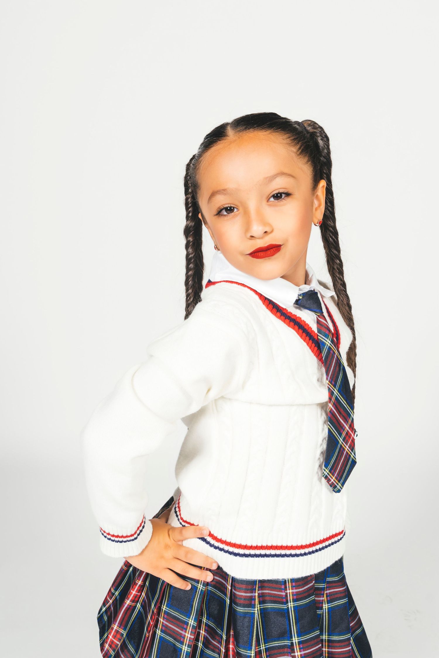 Young girl in plaid school uniform with white sweater, tie, and braids poses confidently against white background.