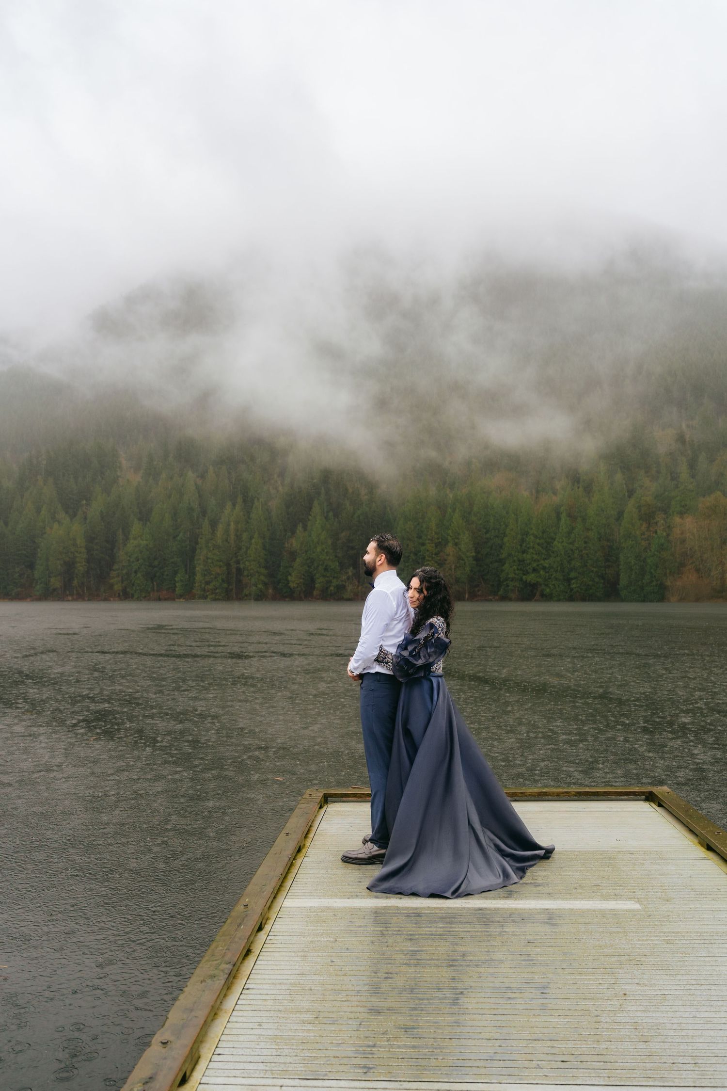 Couple embracing on a misty lake dock surrounded by foggy evergreen mountains in a romantic outdoor setting.