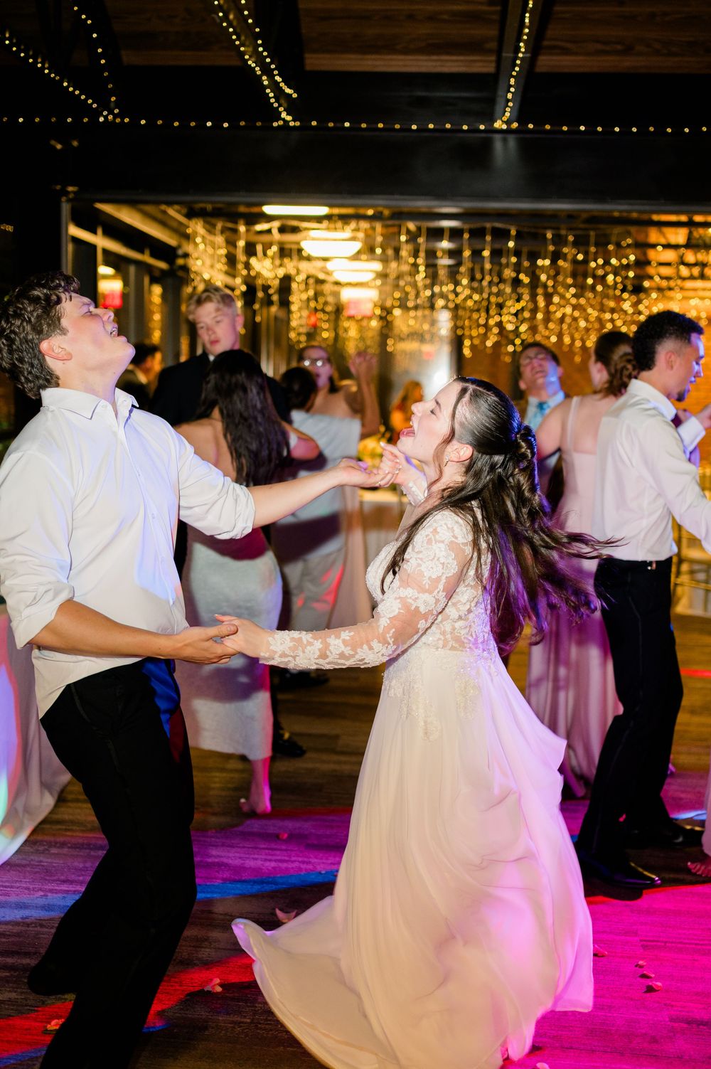 Bride and groom dancing joyfully at wedding reception with guests and fairy lights in background.