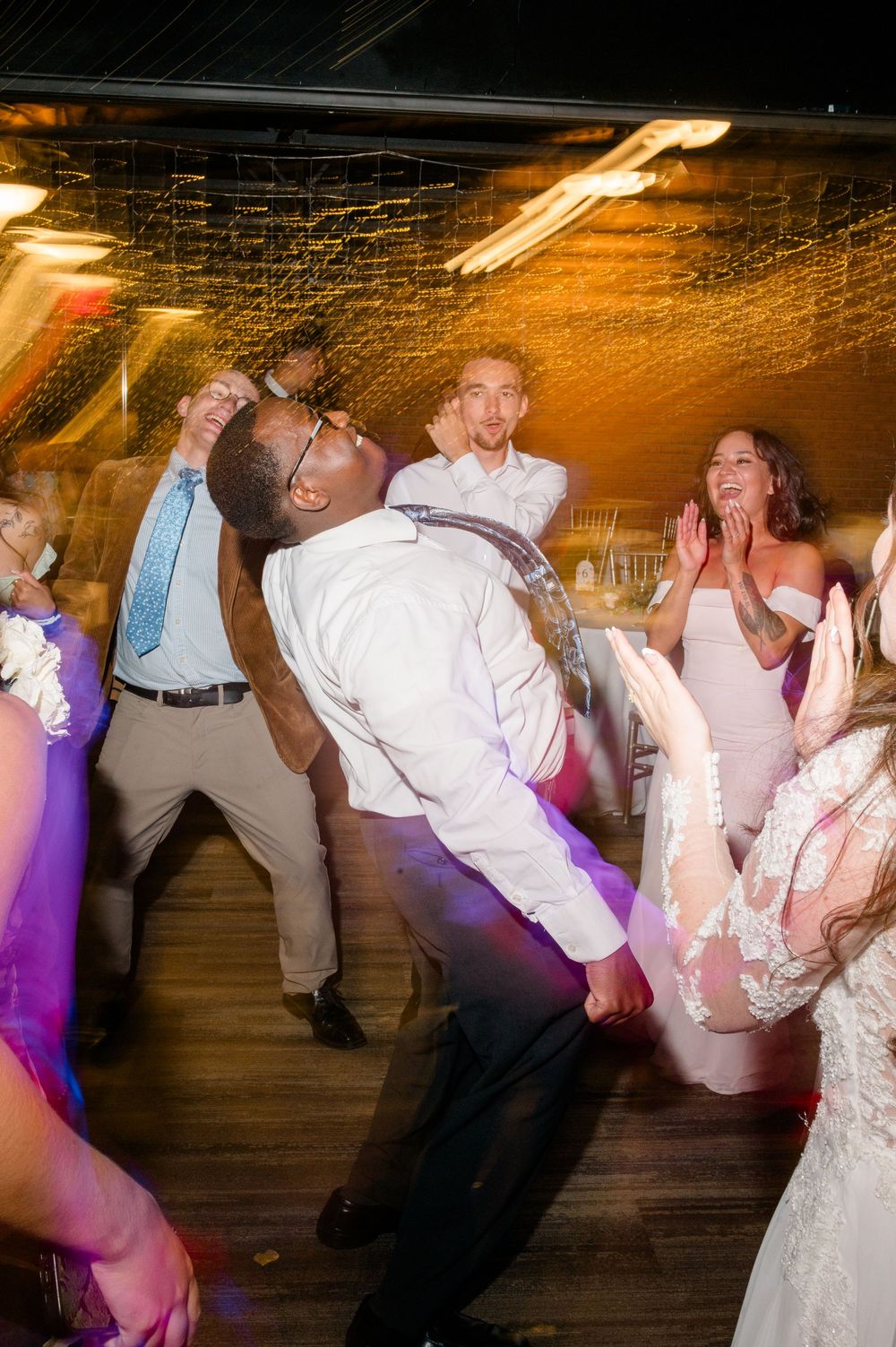 Guests dancing energetically at a wedding reception, with the groom in a white shirt leaning back joyfully on the dance floor.