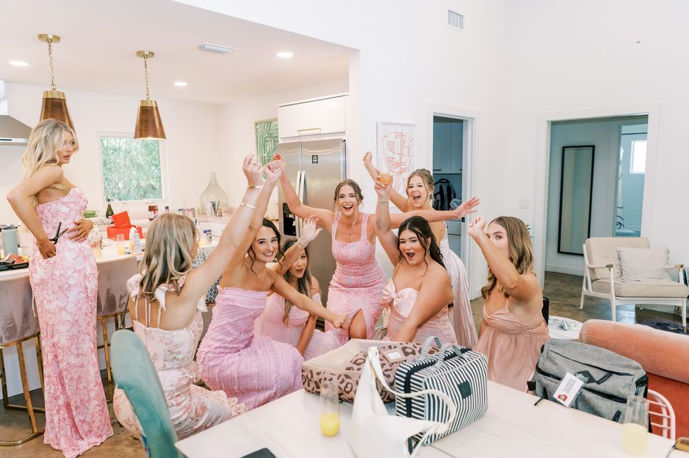 Group of women in pink pajamas celebrating at a bachelorette party, cheering with hands raised in a bright modern home.