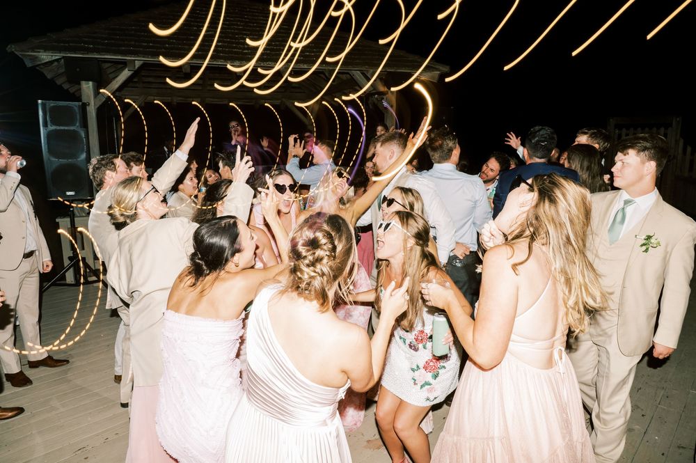 Guests dancing joyfully at a wedding reception under warm string lights, wearing formal attire including pink bridesmaid dresses.