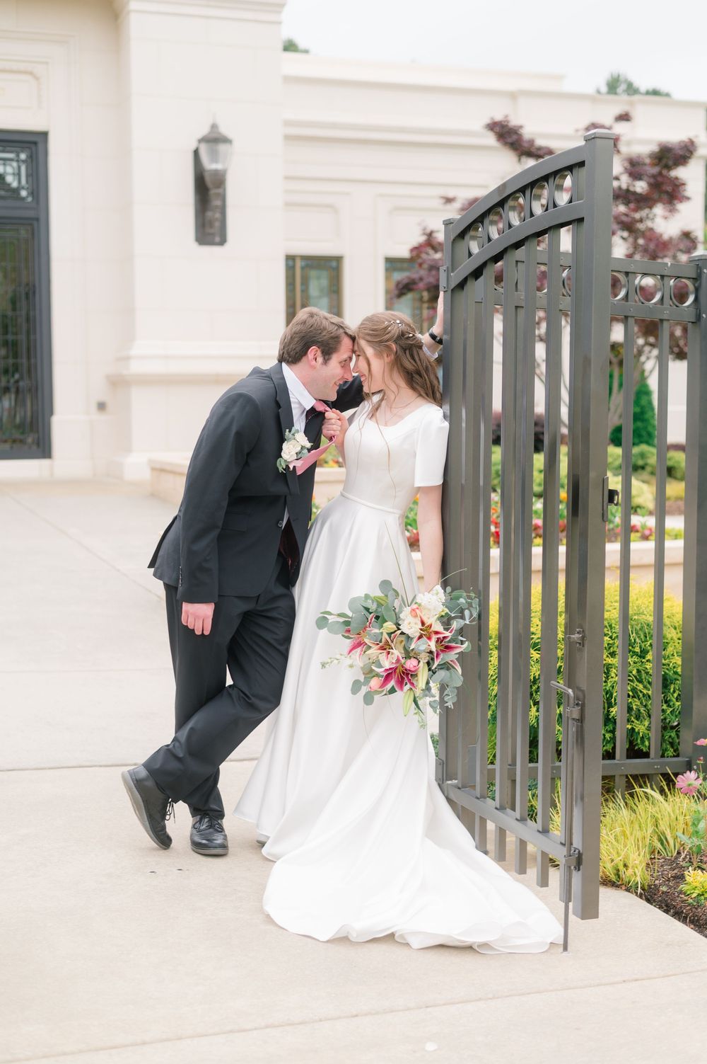 Bride and groom share a romantic moment by an iron gate, bride holding a colorful bouquet in a white gown.
