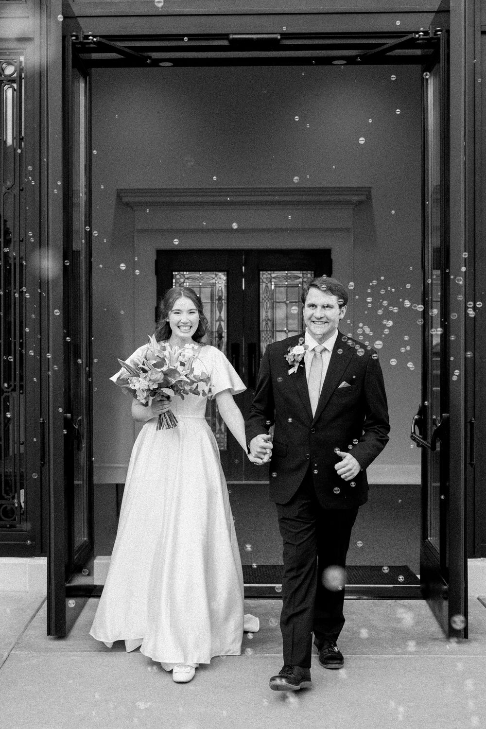 Black and white photo of a smiling bride and groom exiting a building hand in hand, surrounded by bubbles.