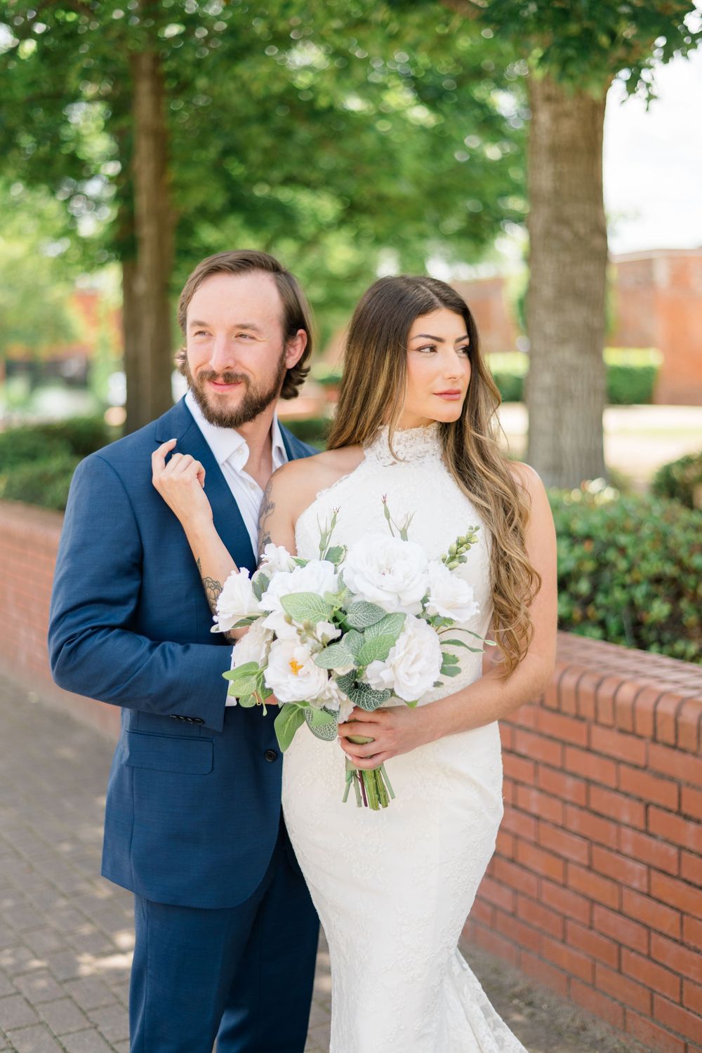 Bride in white halter gown holding white bouquet stands with groom in navy suit outdoors near brick wall and trees.