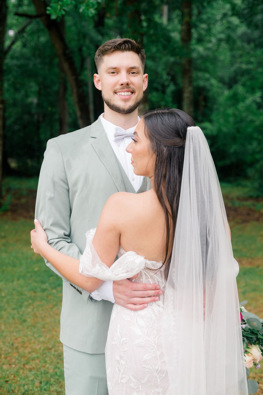 A smiling groom in a sage suit embraces his bride in a white lace gown and veil outdoors.