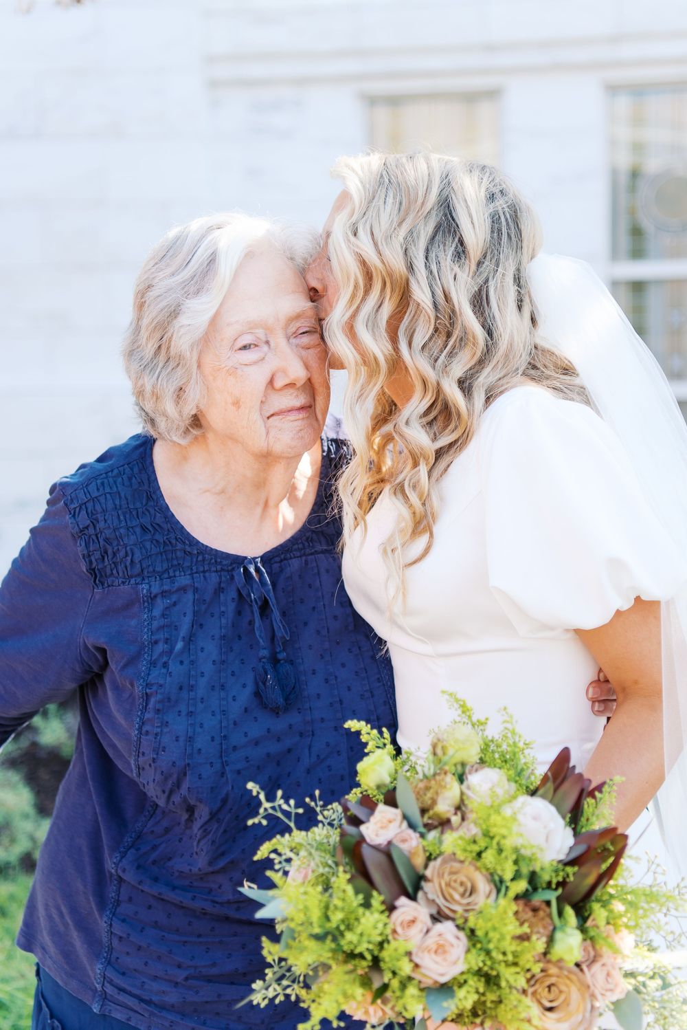 Bride in white gown kisses elderly grandmother on cheek while holding floral bouquet outdoors.