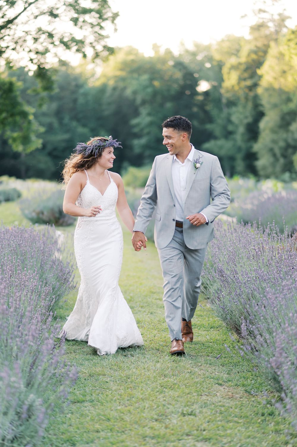 Bride and groom walk hand-in-hand through lavender fields, she in a white gown, he in a gray suit.