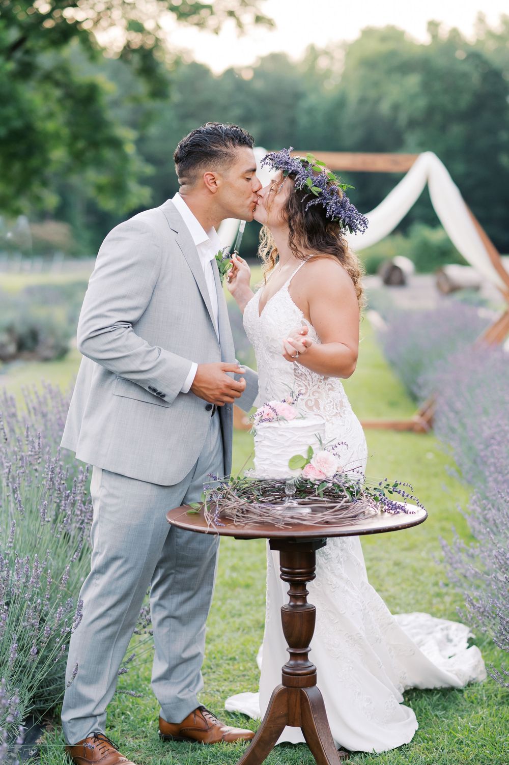 Bride and groom sharing a kiss outdoors in a lavender field, bride wearing floral crown, groom in light gray suit.