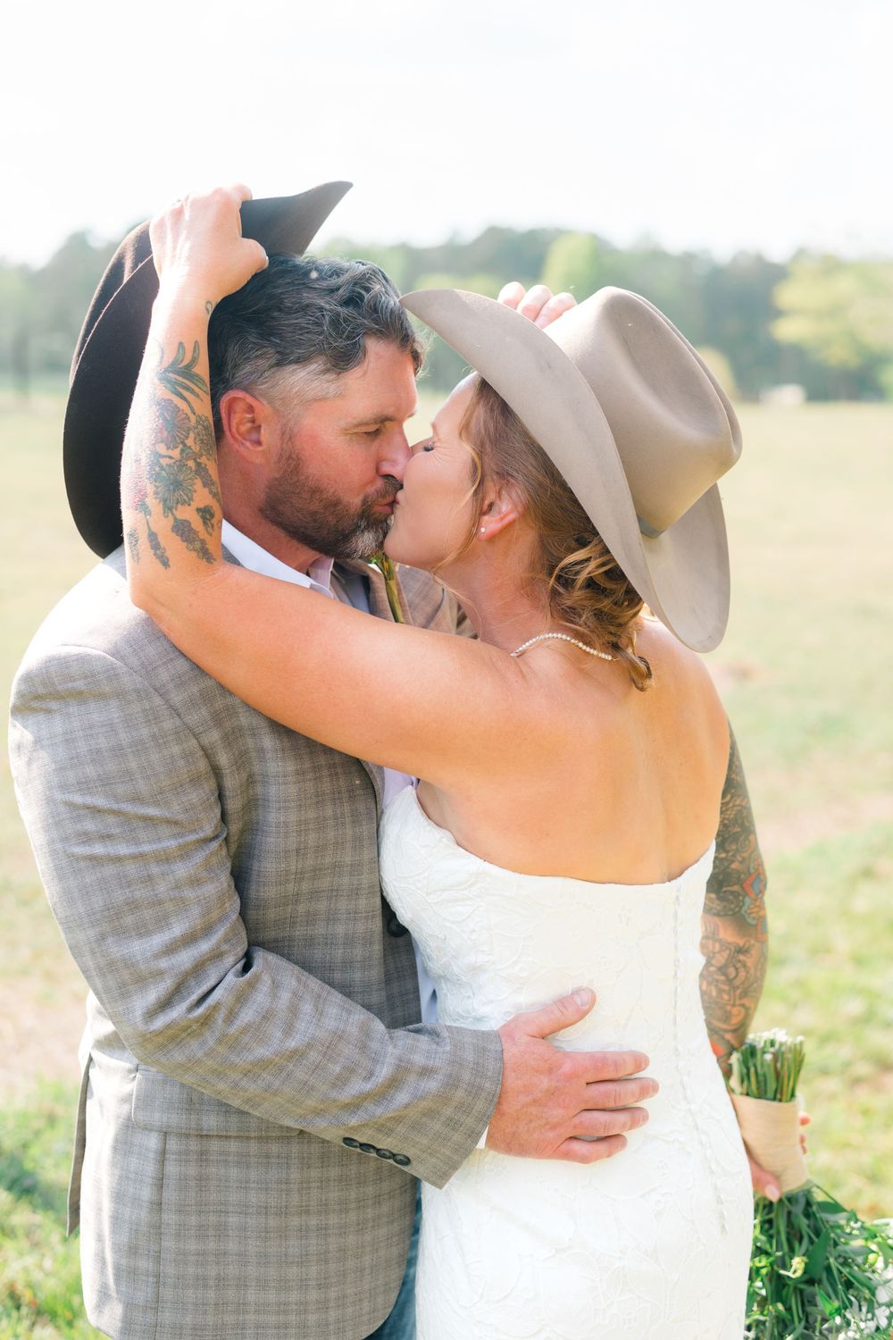 A tattooed bride and groom share a kiss in a field, both wearing cowboy hats, with the bride in white and groom in gray.