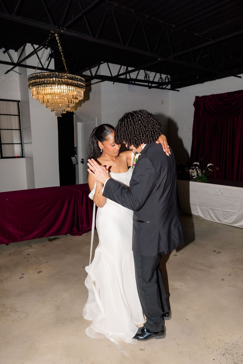 Bride and groom share their first dance under a chandelier in an elegant venue with burgundy drapes.
