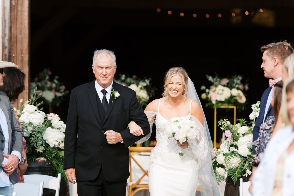 Bride walks down the aisle with her father in a barn wedding venue surrounded by white floral arrangements.