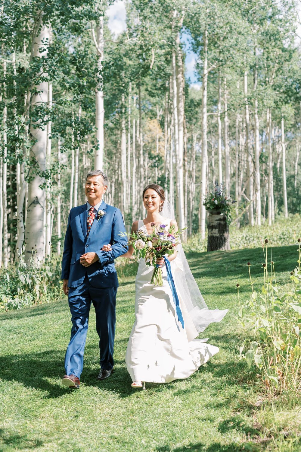 Bride in white gown with bouquet walks with father through aspen grove on sunny wedding day.