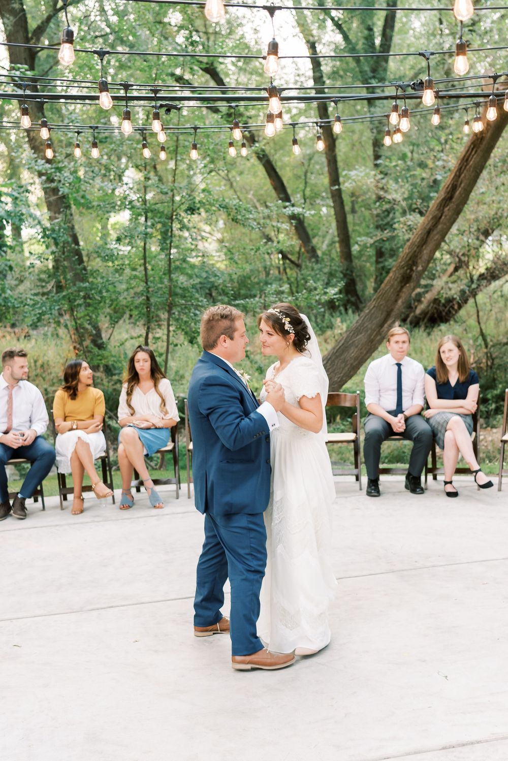 Bride and groom share first dance outdoors under string lights with guests seated nearby at a romantic woodland wedding reception.