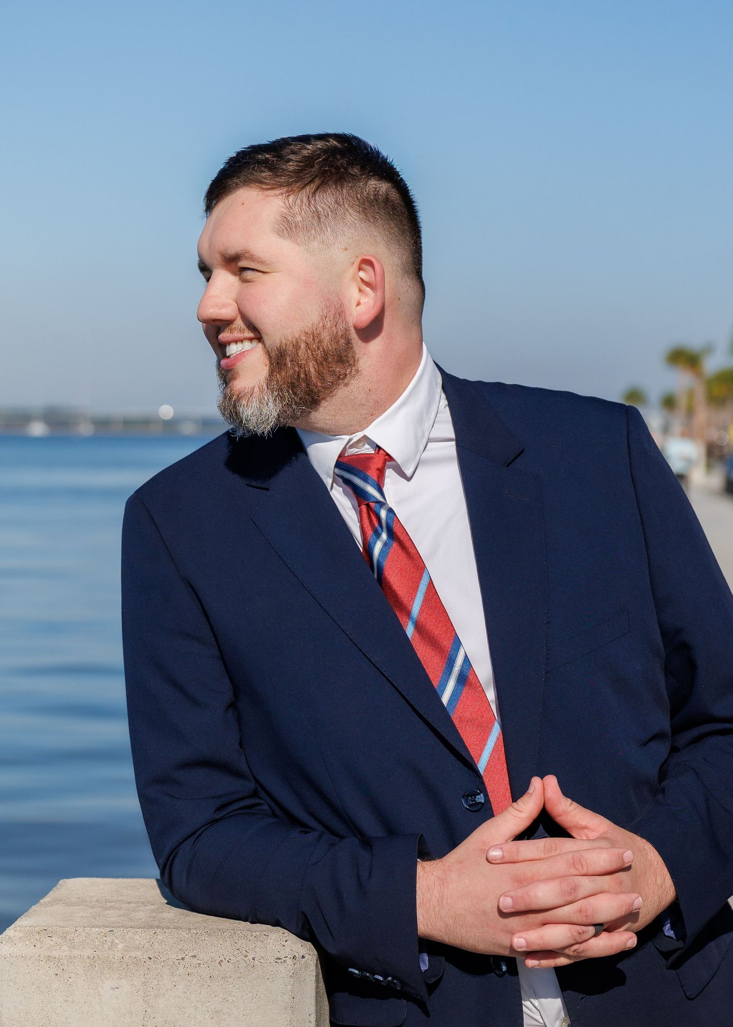 Man in navy suit and red plaid tie smiling outdoors by waterfront on sunny day.