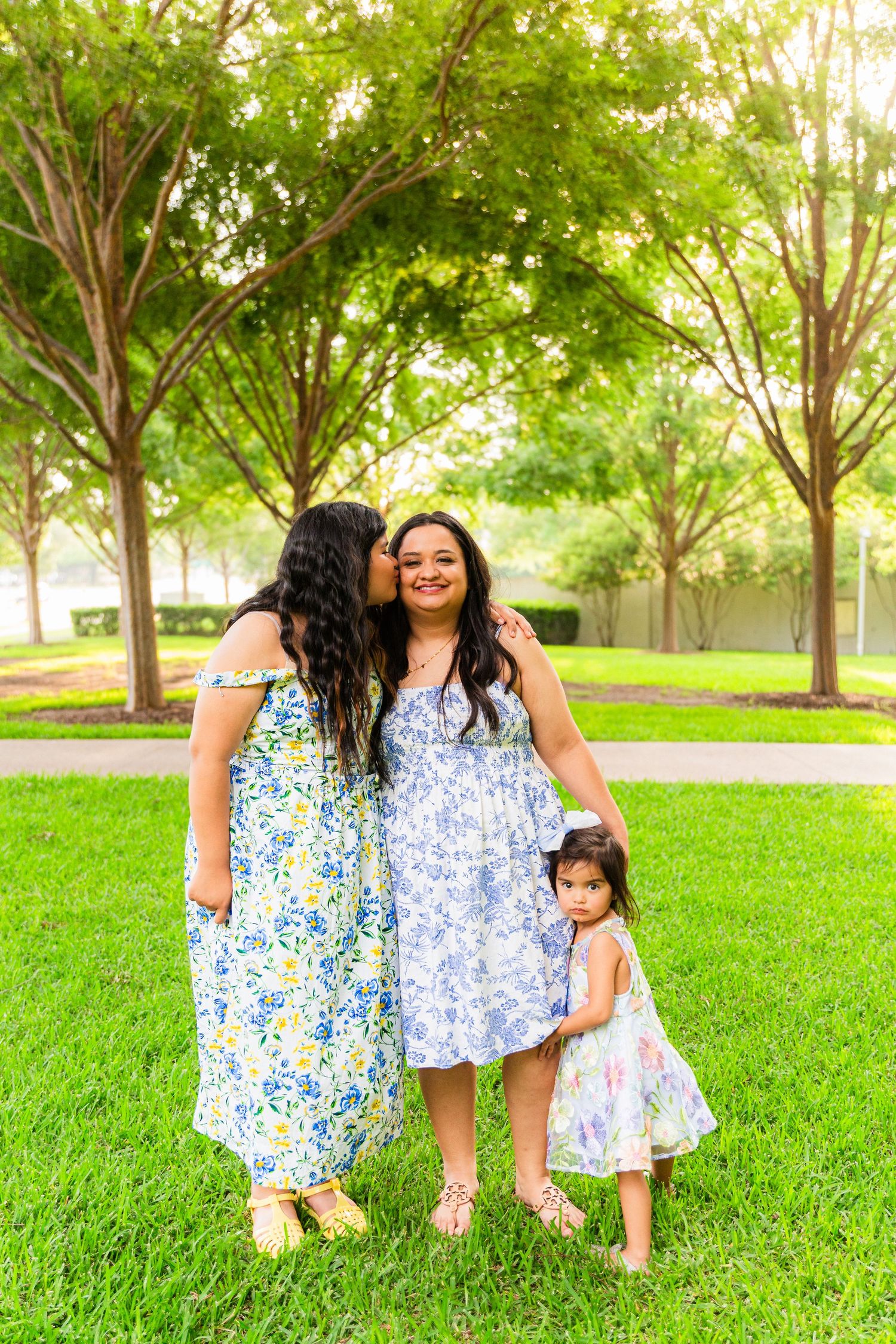Three generations of women in floral dresses pose together on green grass in a sunny park setting.