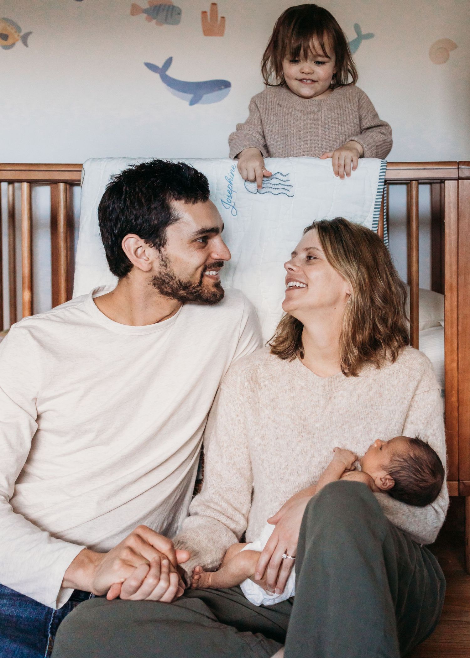 Happy family with newborn baby and toddler sitting by a crib in a nursery with ocean animal wall decals.