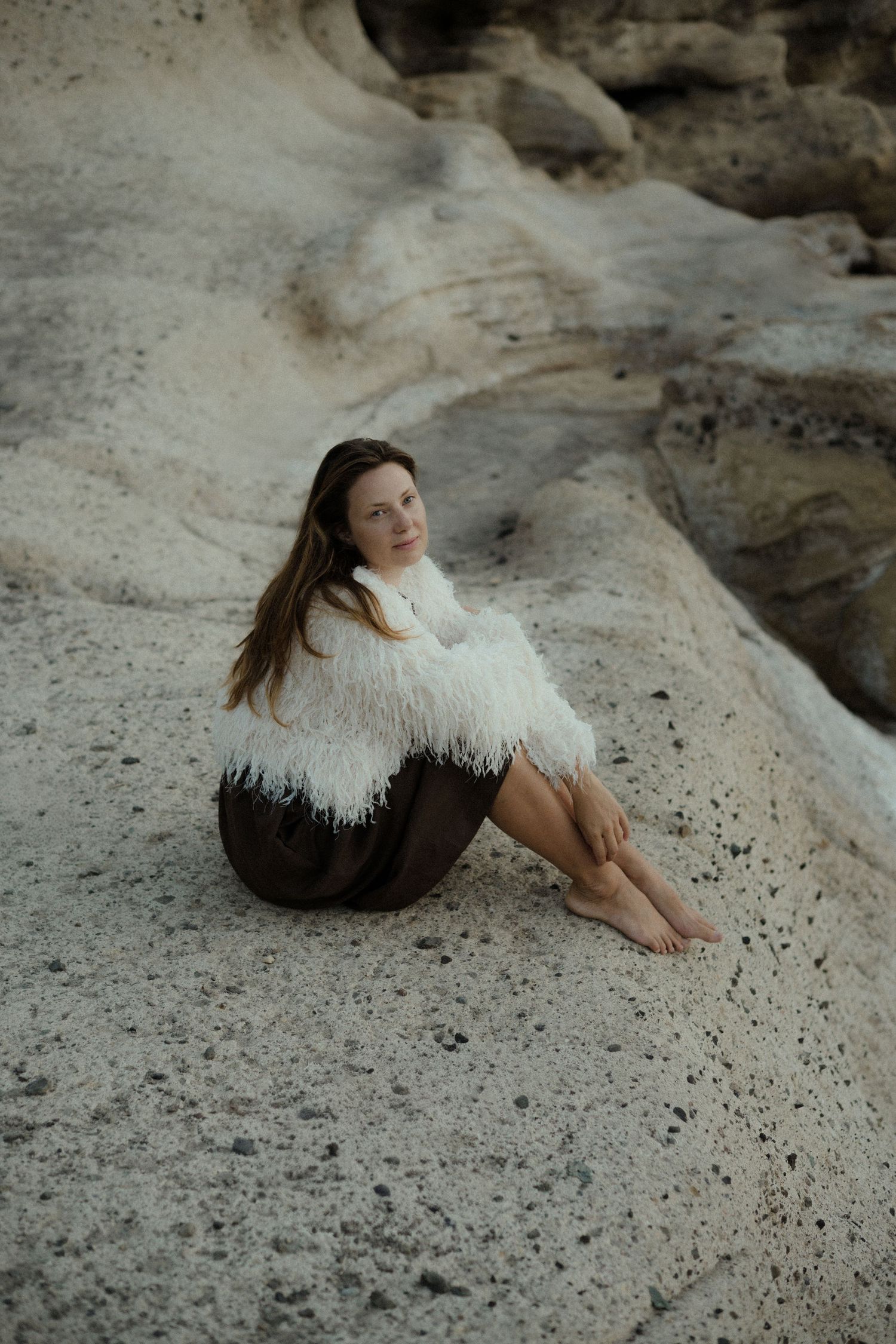 A barefoot woman in a white fluffy jacket and brown skirt sits on sandy rock formations outdoors.