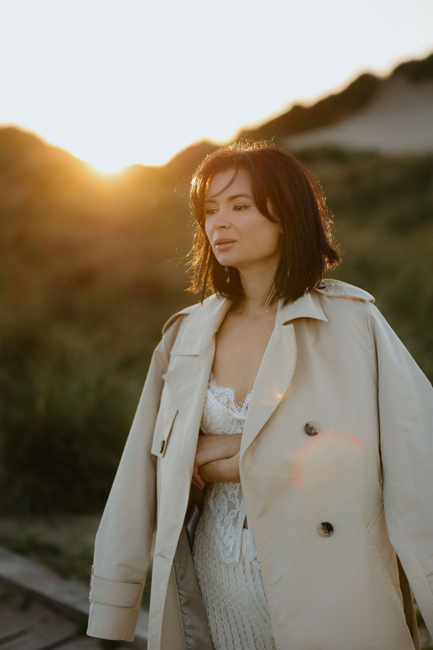 Woman in beige trench coat standing outdoors at golden sunset with hills in background.