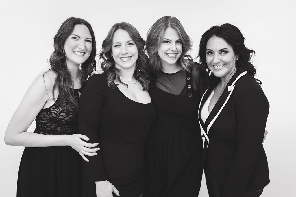 Four women in black outfits smiling together in a professional black and white portrait photo.