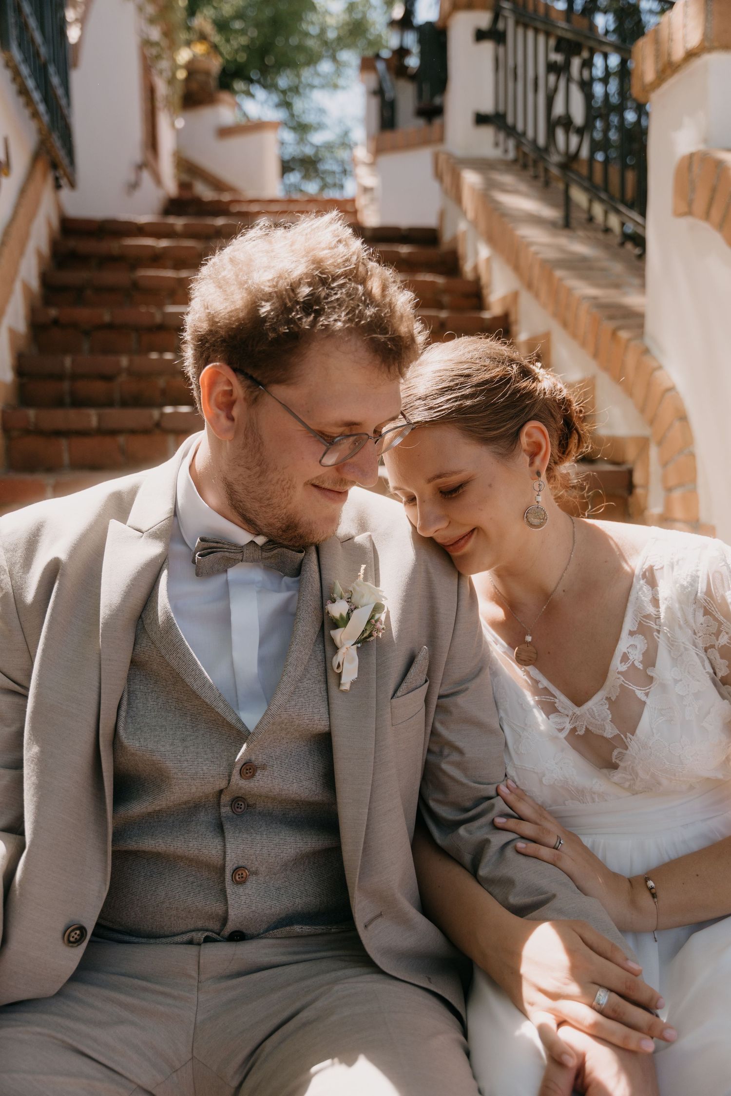 A newlywed couple in wedding attire sit closely together on outdoor steps, sharing a tender romantic moment in warm sunlight.