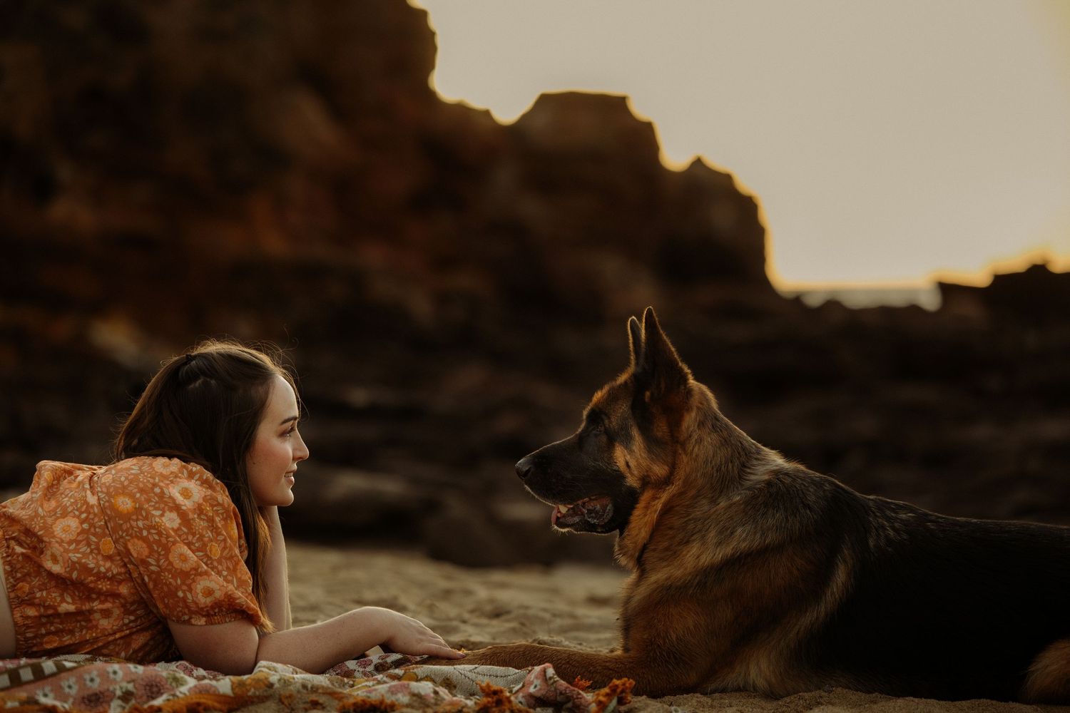 A young woman in an orange floral dress lies face-to-face with a German Shepherd dog on a rocky beach at sunset.