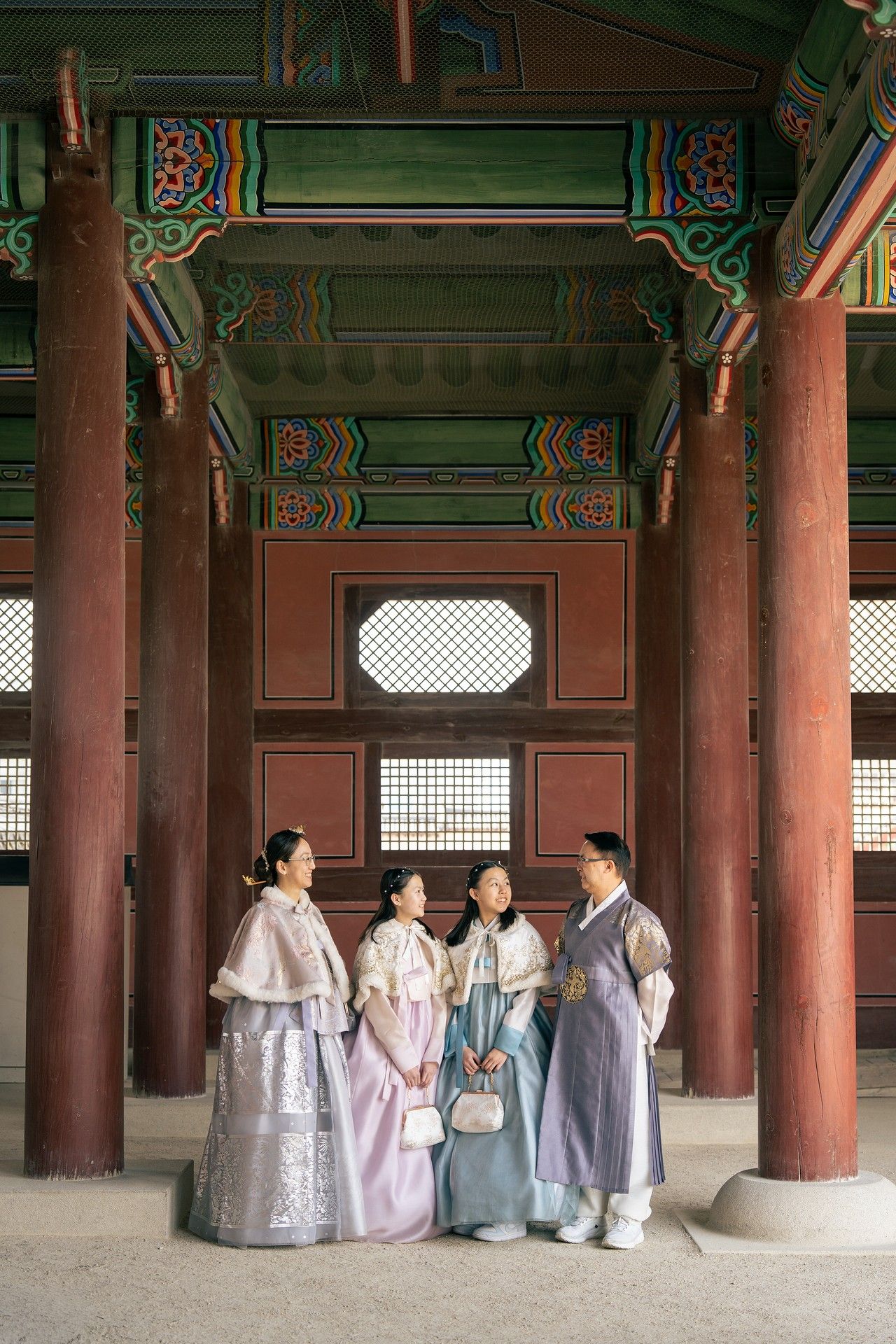 Four people wearing traditional Korean hanbok stand together at a historic palace pavilion with colorful painted wooden architecture.