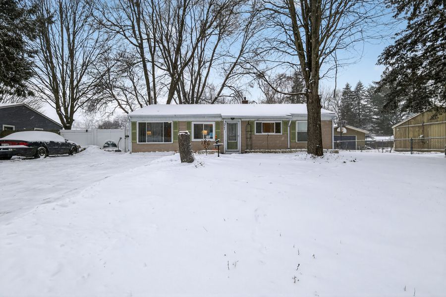 Snow-covered ranch-style home with green siding, large trees, and a spacious front yard in winter.