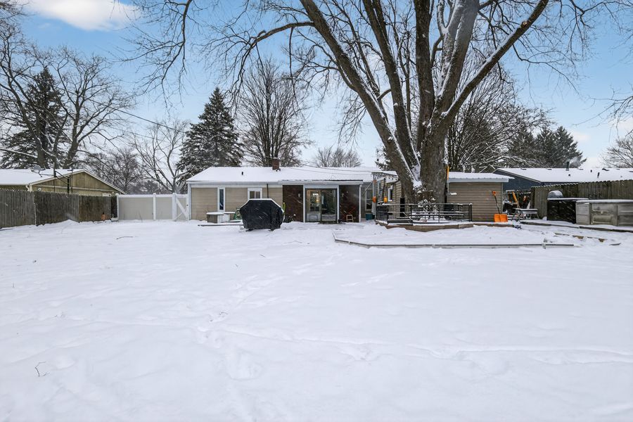 Snow-covered backyard with large bare tree, single-story home, BBQ grill, and wooden privacy fence.
