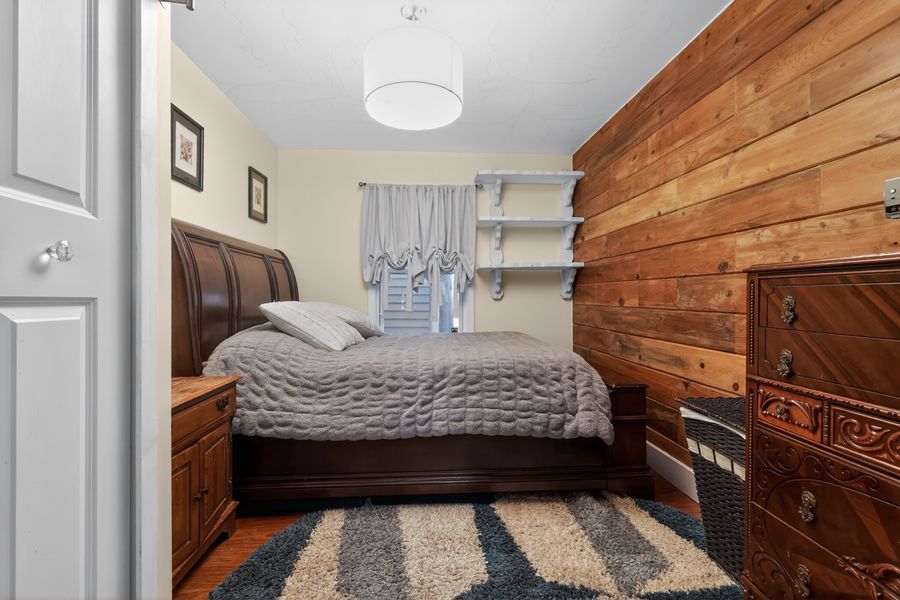 Cozy bedroom with wood-paneled accent wall, dark wood furniture, gray bedding, striped rug, and a bright window with curtains.
