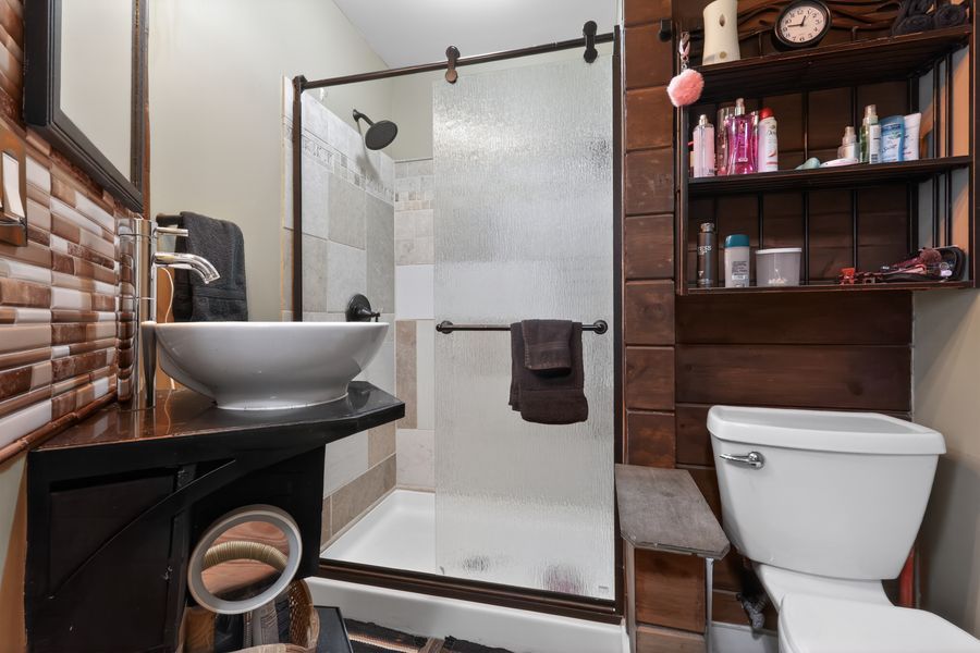 Modern bathroom with vessel sink, sliding glass shower, brick accent wall, wooden shelving, and white toilet.