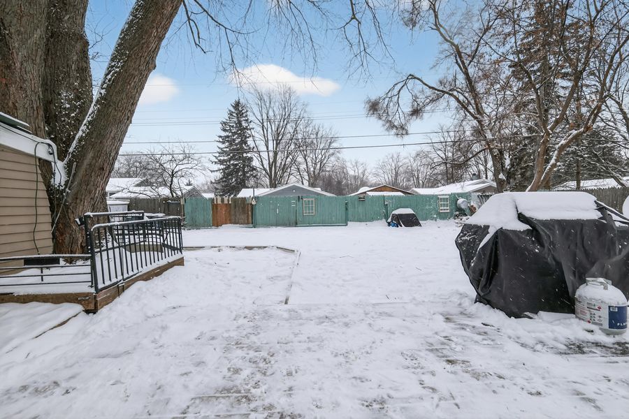 Winter backyard scene with deep snow, large trees, green storage sheds, and a covered item near the fence.