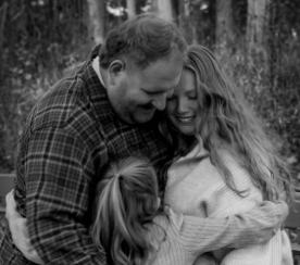 Black and white photo of a loving family of three sharing a warm embrace outdoors.