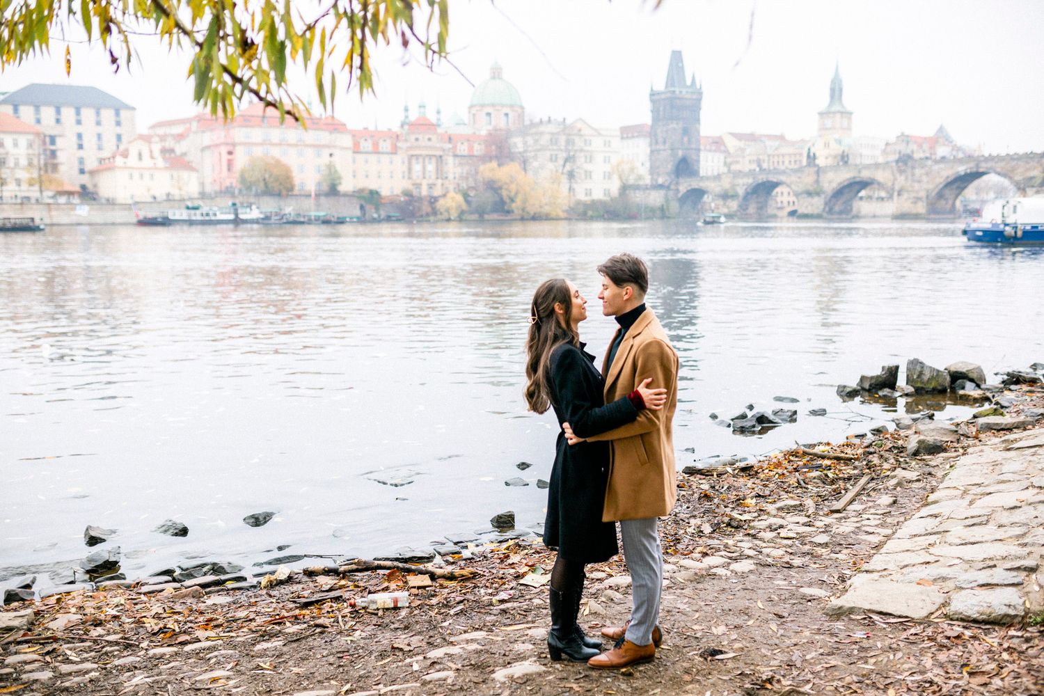 Romantic couple embracing by Prague's Vltava River with the Charles Bridge in background.