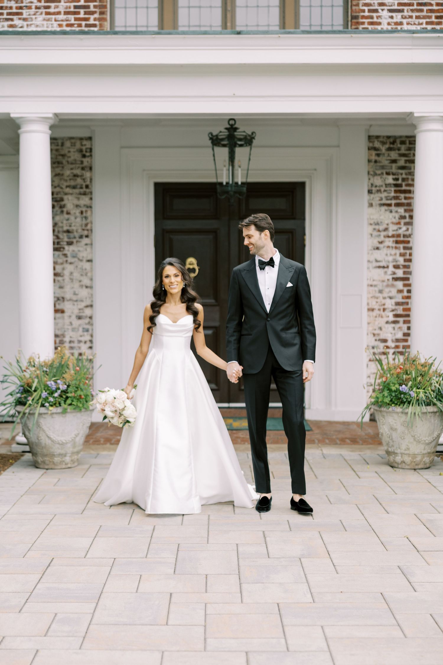 Bride in white ball gown and groom in black tuxedo hold hands outside an elegant brick estate.