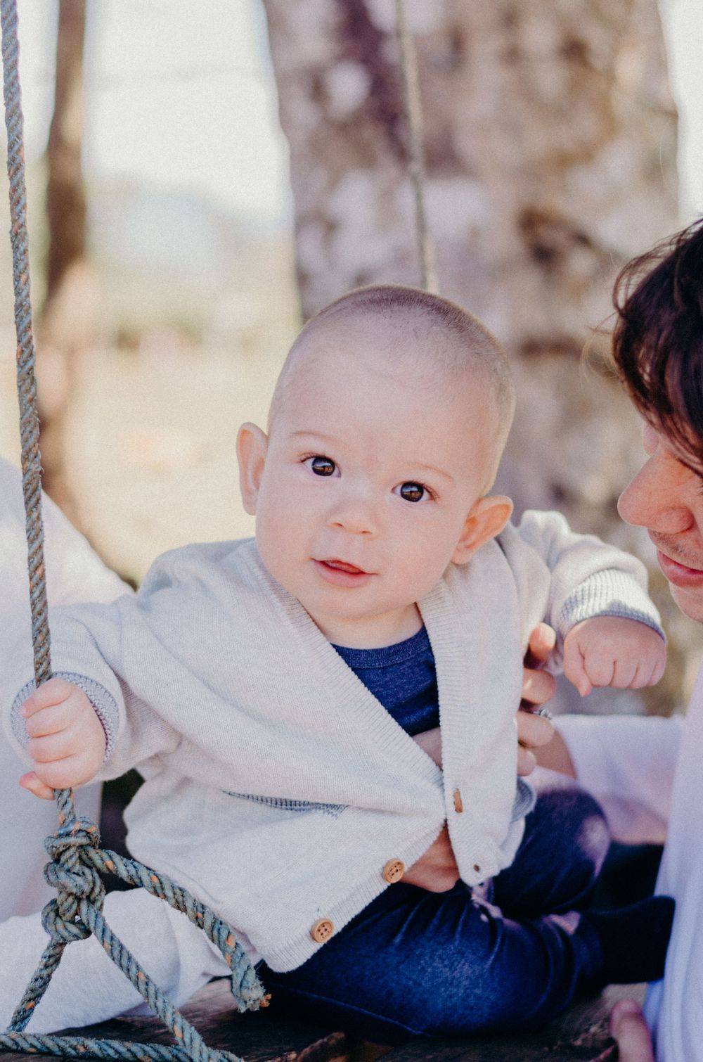 Baby on wooden swing wears light gray cardigan against blurred background.