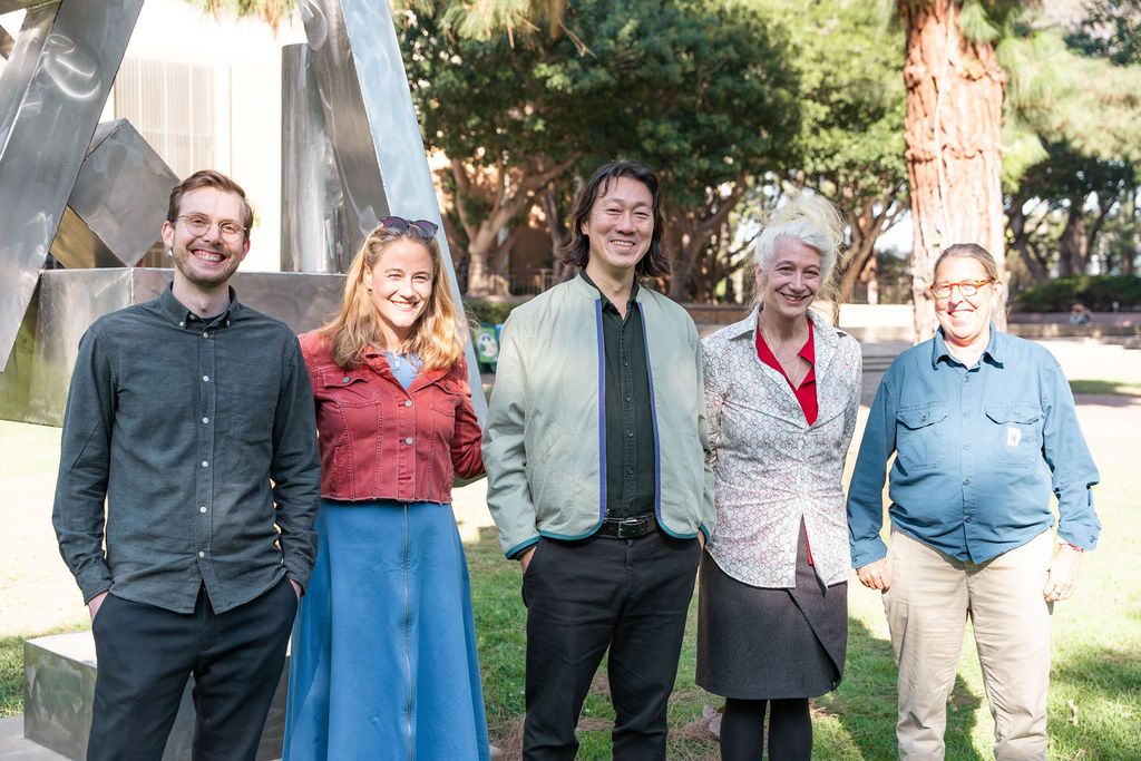 Five people smiling and posing outdoors near a modern sculpture and palm trees on a sunny day.