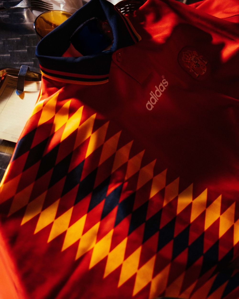 Adidas Spain retro football shirt with diamond pattern in red, yellow, and black displayed in warm sunlight.