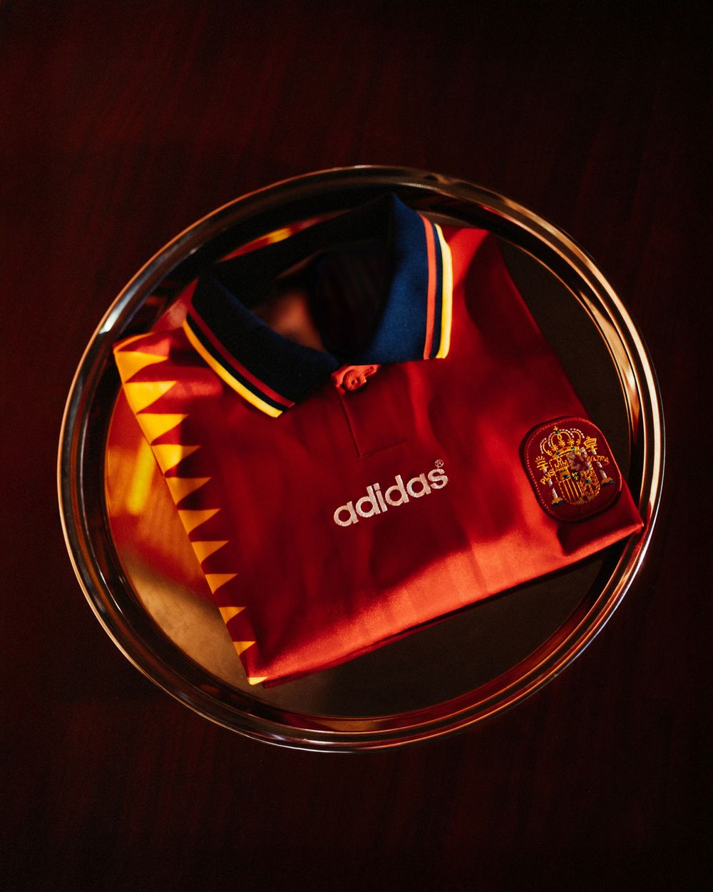 Folded Adidas Spain retro red football shirt with crest displayed in a glass bowl on dark background.