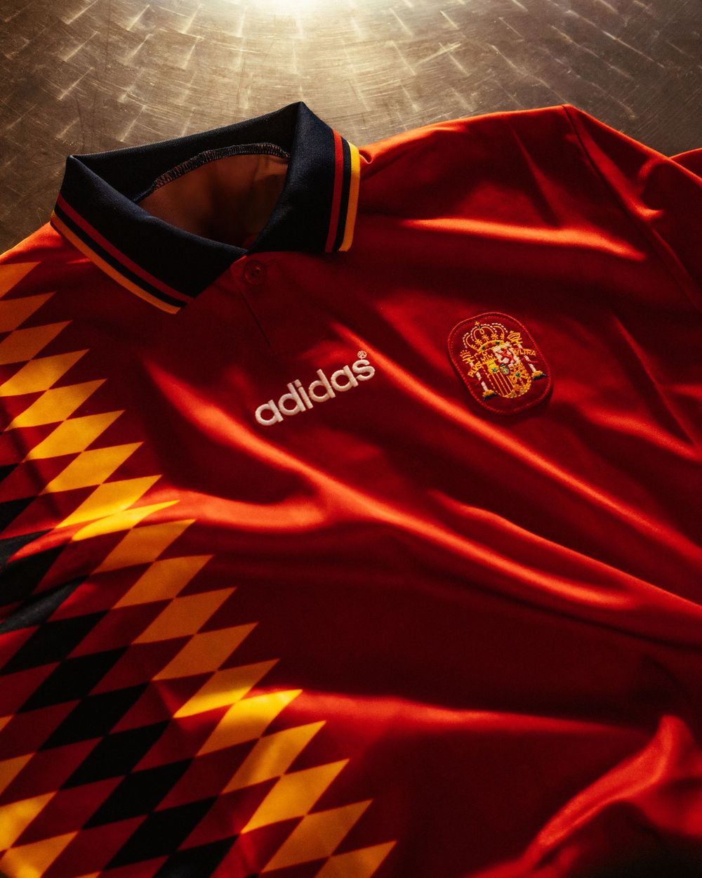 Adidas Spain retro red football jersey with diamond pattern and navy collar displayed on a wooden surface.