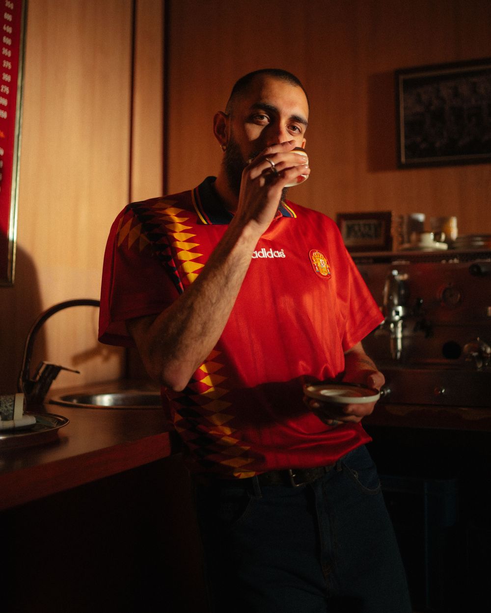 Man in red Adidas soccer jersey holding a cup in a warmly lit kitchen.