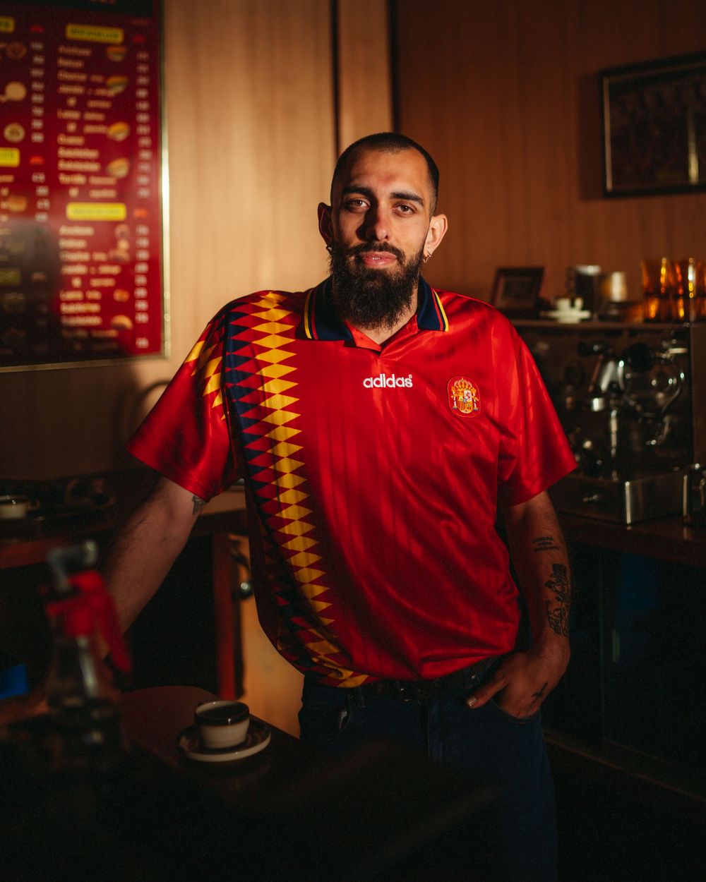 Man with beard wearing a red Adidas Spain football jersey poses confidently in a dimly lit cafe bar setting.