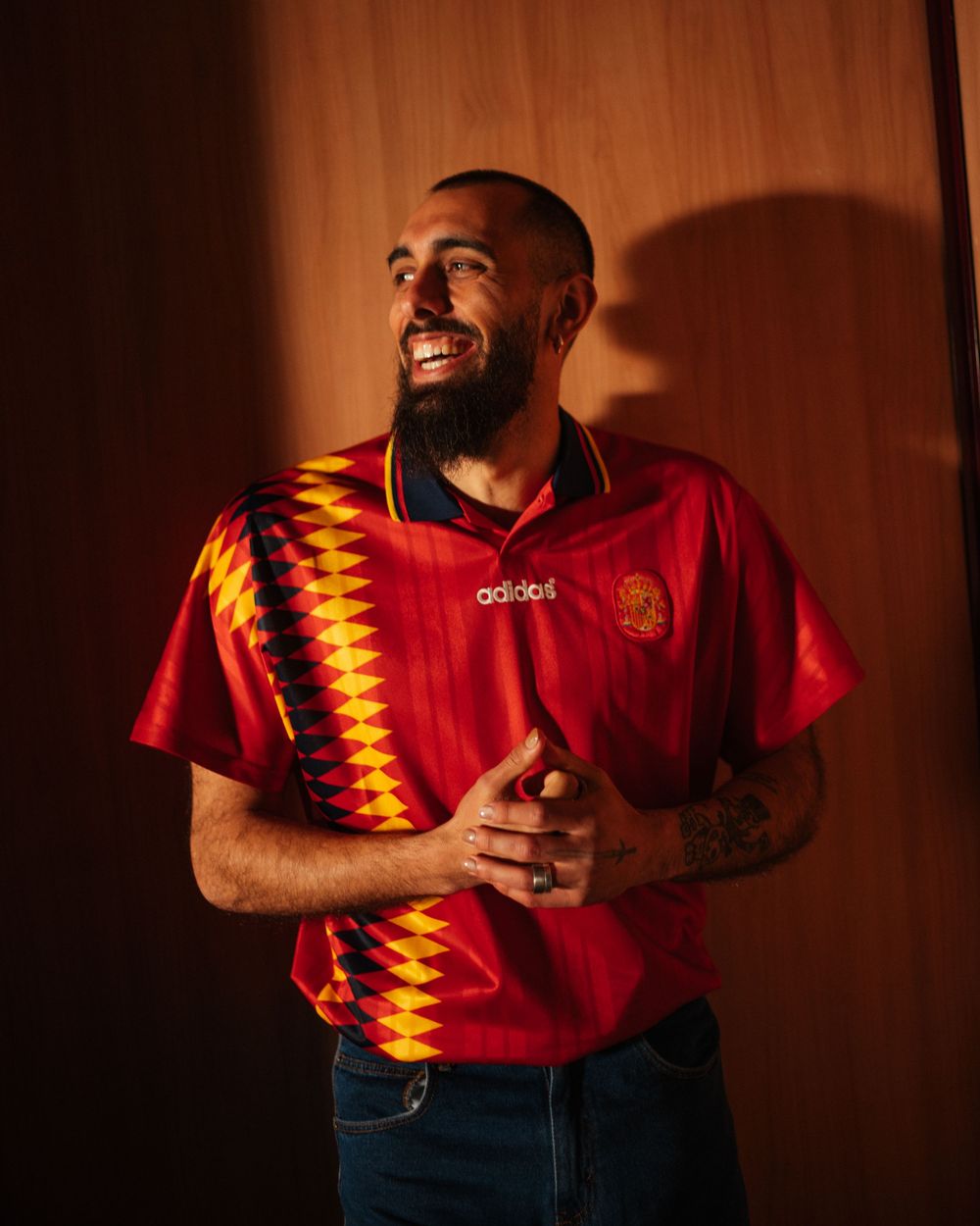 Smiling man with beard wearing a retro red Adidas Spain football jersey with diamond pattern, posing indoors.