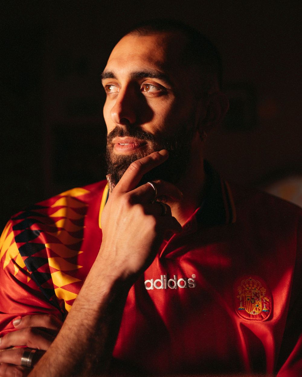 Man with beard wearing red Adidas Spain football jersey, thoughtfully touching his chin in dramatic lighting.