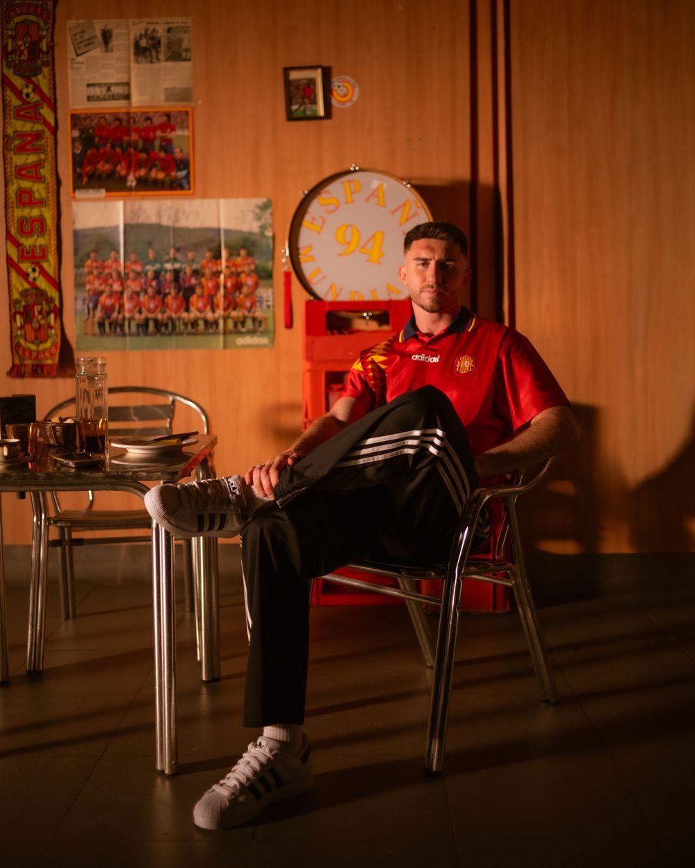 Young man in red football jersey sits relaxed on metal chair in retro room with sports posters and memorabilia.